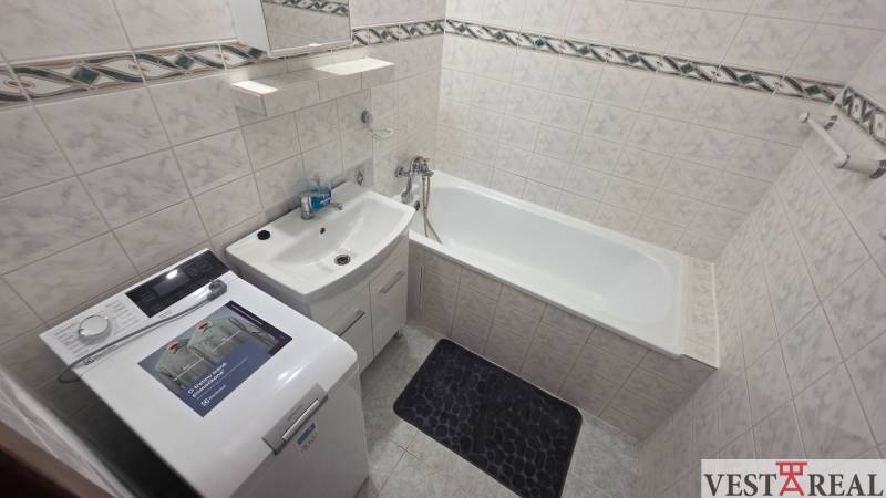 Fürdőszoba káddal és mosógéppel egy egyszobás lakásban, világos csempék és tárolóhely.