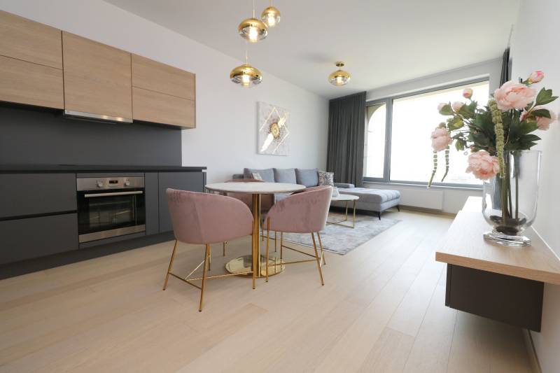 Nappali konyhával, fa mintázatú padlóval, világos kanapéval, étkezőasztallal, virágokkal.