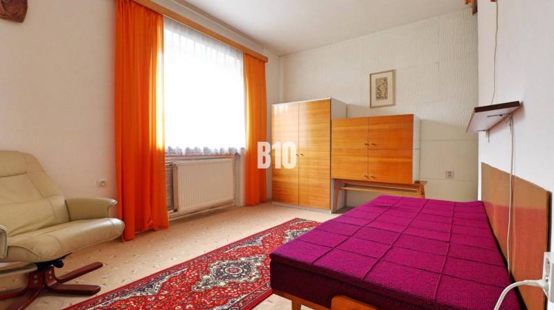 Szoba szőnyegpadlóval, krémszínű karosszékkel, színes ággyal és szekrénnyel egy családi házban.