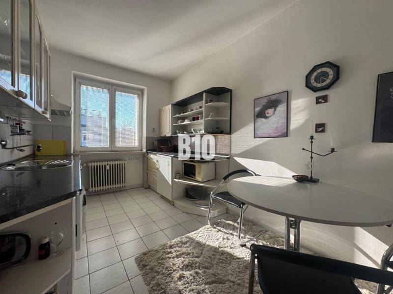 Konyha egy kétszobás lakásban fehér szekrényekkel, ablakkal és étkezőasztallal.