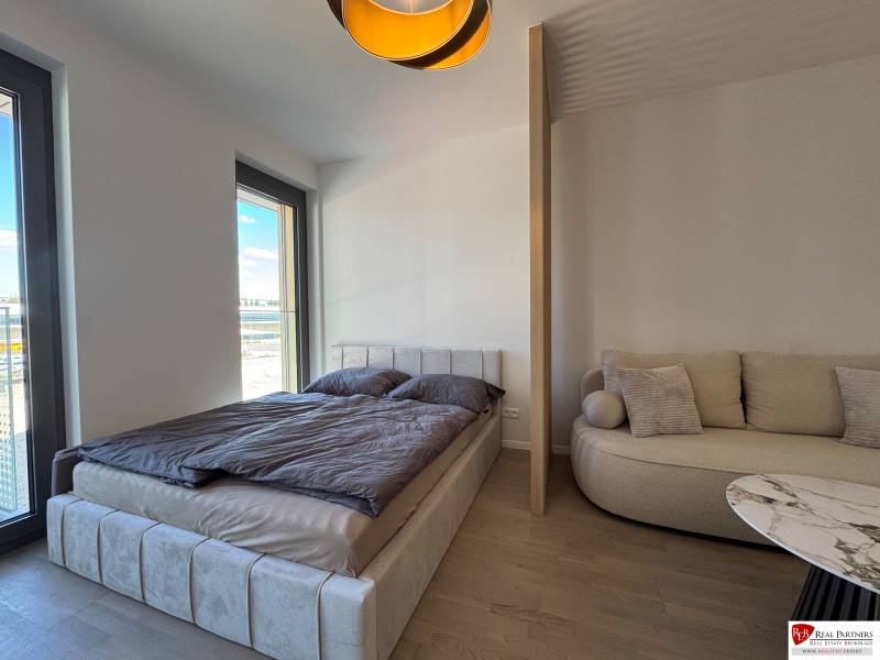 Hálószoba egyszobás lakásban ággyal, kanapéval és fa mintázatú padlóval.