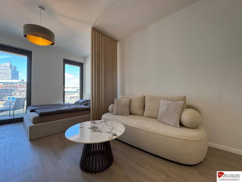 Nappali kanapéval, dohányzóasztallal és ággyal egy egyszobás lakásban, fa dekor padlóval.