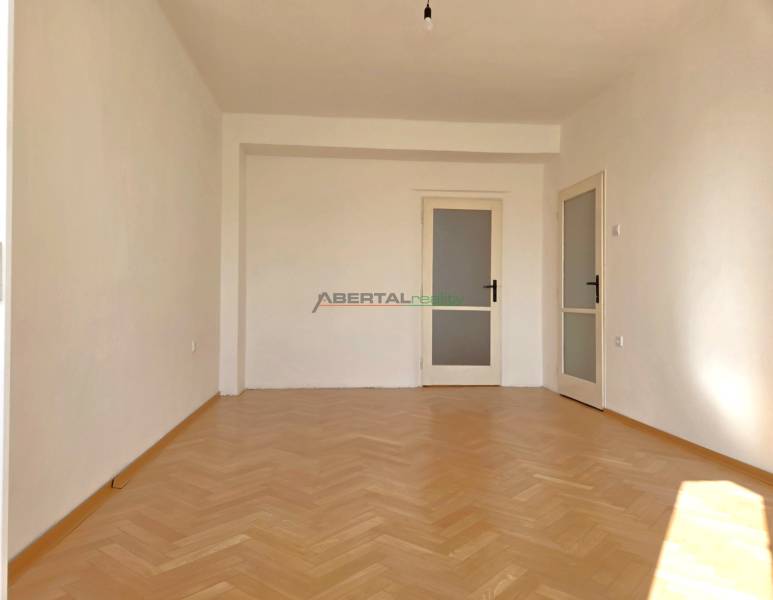 Egy szoba a háromszobás lakásban fa dekorációjú padlóval és fehér falakkal rendelkezik.