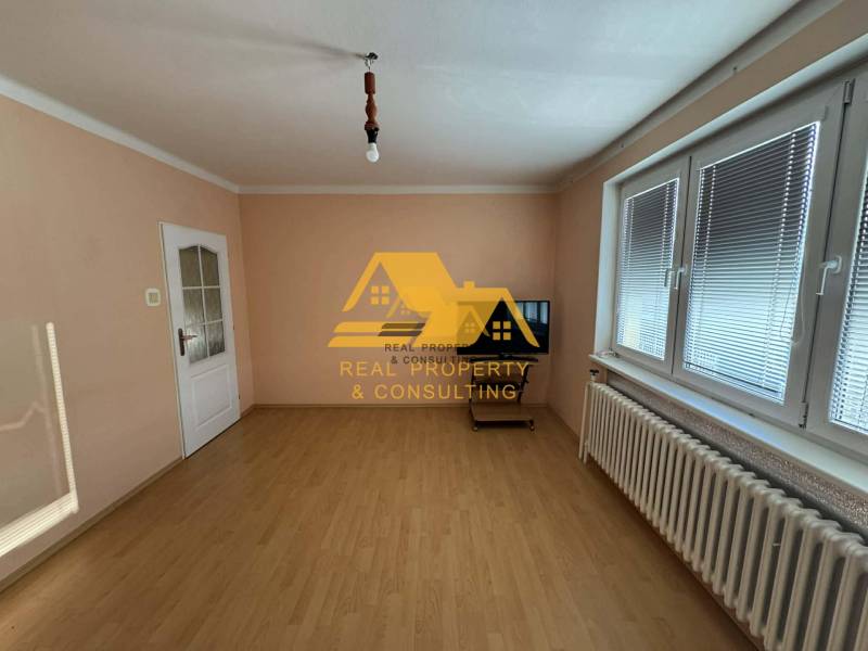 Egy családi ház szobája fehér falakkal, fa mintázatú padlóval és redőnyös ablakkal.