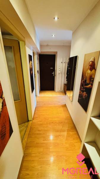 Keskeny folyosó fa mintázatú padlóval egy háromszobás lakásban, képekkel és tükörrel.