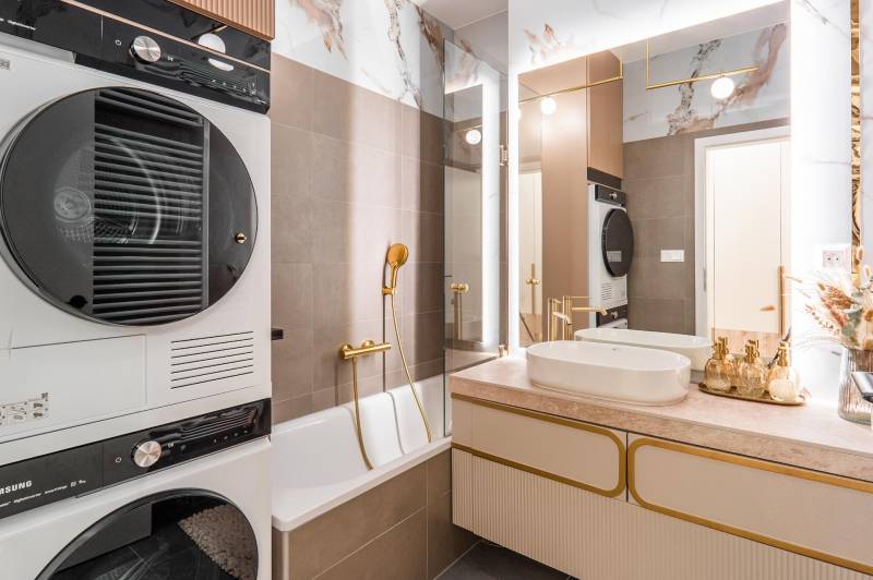 Fürdőszoba egy háromszobás lakásban káddal, mosdóval, szárítóval és elegáns arany kiegészítőkkel.