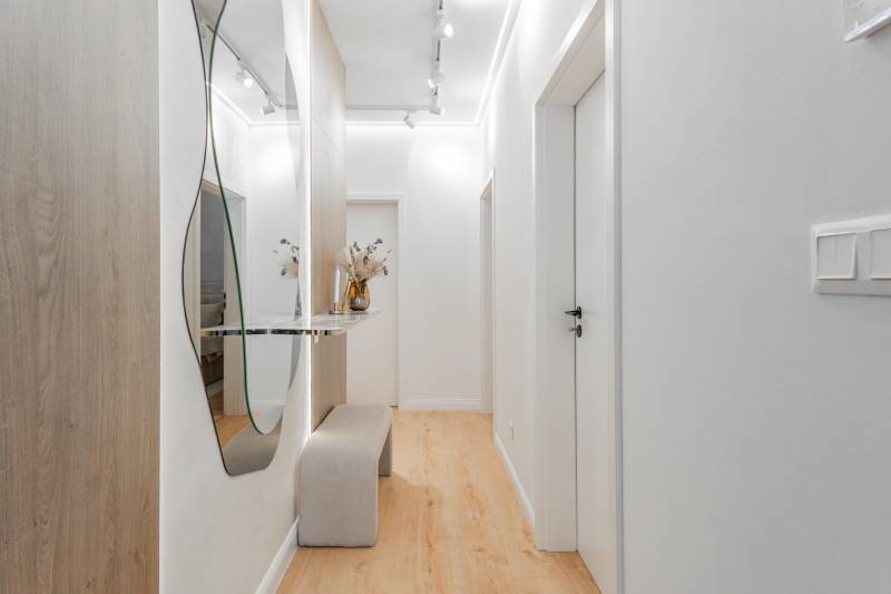 Világos előszoba tükörrel, paddal és fa dekorációjú padlóval egy háromszobás lakásban.