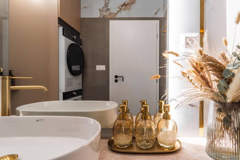 Fürdőszoba mosdóval, arany adagolókkal és dekoratív természetes díszítéssel egy háromszobás lakásban.