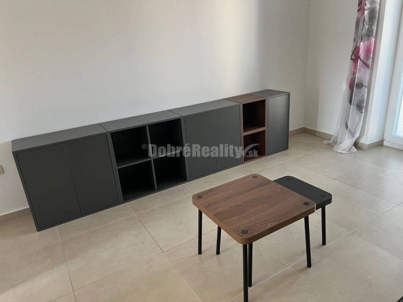 Nappali egy kétszobás lakásban, járólappal, asztallal és sötét szekrénnyel.