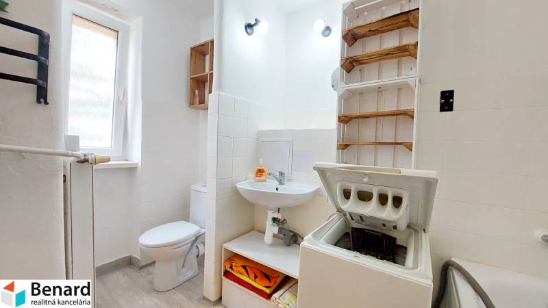 Fürdőszoba egy kétszobás lakásban mosógéppel, szaniterrel és törölközőtartó polccal.