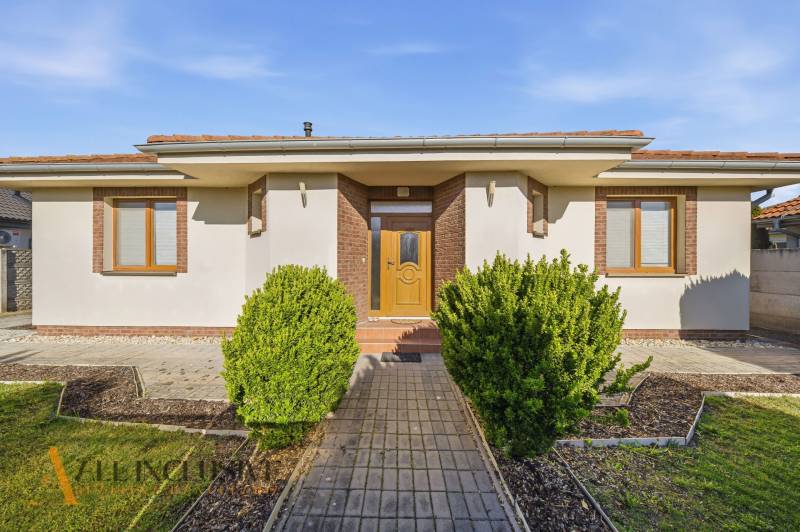 Családi ház Kútnikokban bokros ültetéssel, fehér homlokzattal és barna kiegészítőkkel.