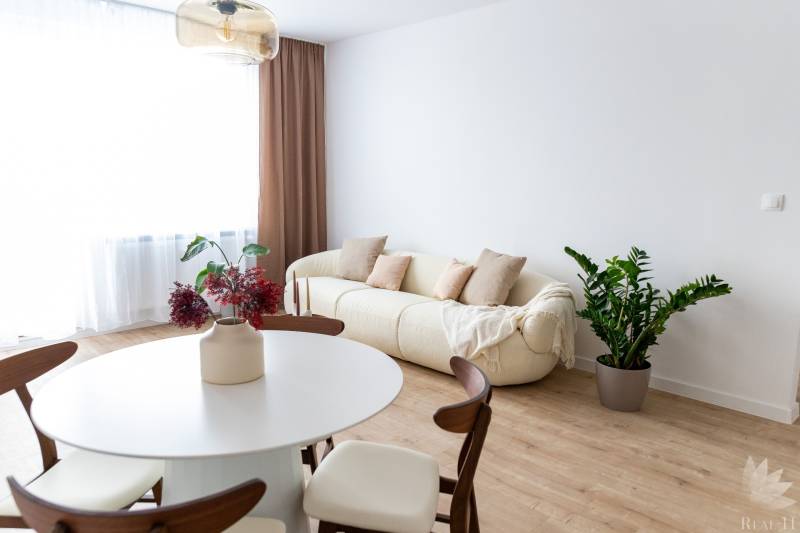 3 szobás lakás belső tere krémszínű kanapéval, növénnyel és asztallal fa dekorációjú padlón.