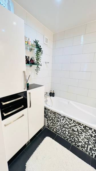 Fürdőszoba egy háromszobás lakásban fehér káddal, modern csempékkel és tárolóhellyel.