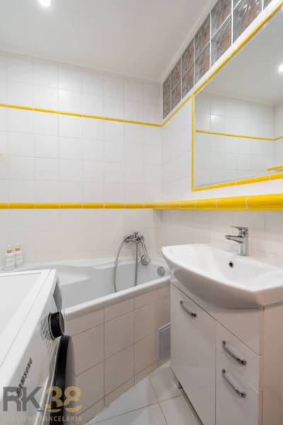 A nyaraló apartman fürdőszobája káddal, mosdóval, mosógéppel és fehér csempékkel.