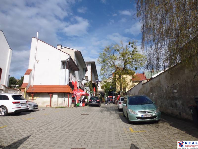 A főutcán Eperjesen autók parkolnak és gyalogosok vannak a kövezett területen.