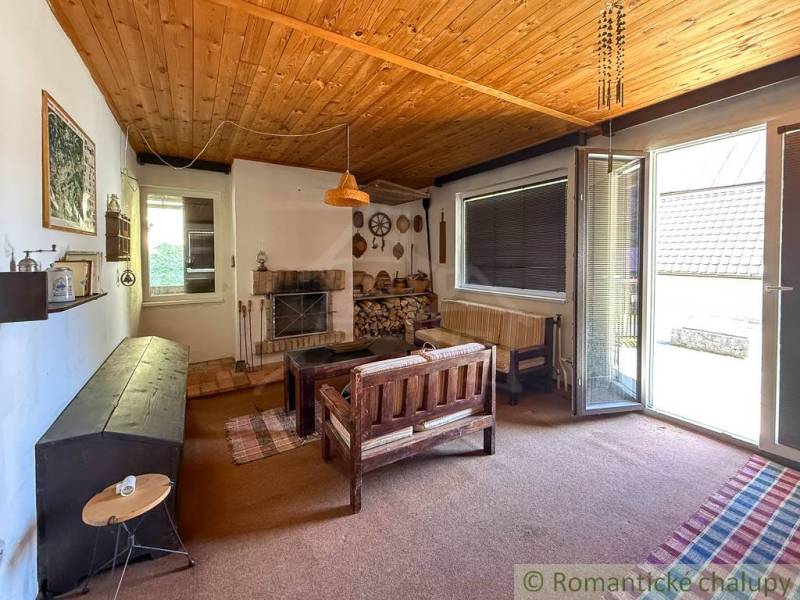 Egy faháznak a belső tere, fából készült mennyezettel, ülőgarnitúrával és kandallóval, kilátással a kertre.