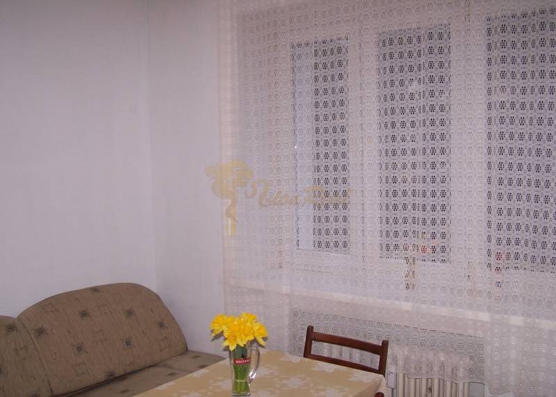 Szoba kanapéval, asztallal, székkel és függönnyel, az asztalon egy váza virágokkal.