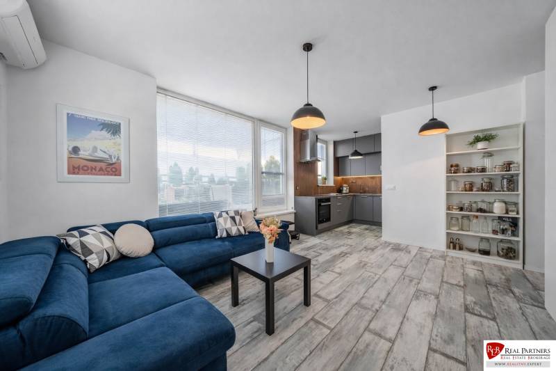 Nappali fa hatású padlóval, kék kanapéval és konyhasarokkal egy kétszobás lakásban.