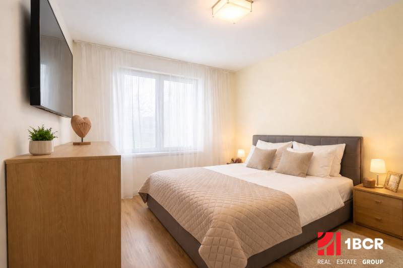 Hálószoba egy háromszobás lakásban szürke ággyal, fa mintázatú padlóval és falra szerelt TV-vel.