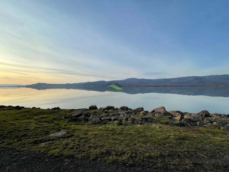 Nyugodt vízfelület, füves part és hegyek Vinné környékén. Lakóterületek.