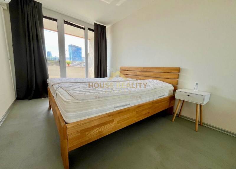 Hálószoba fa ággyal, éjjeliszekrénnyel és erkéllyel egy háromszobás lakásban.