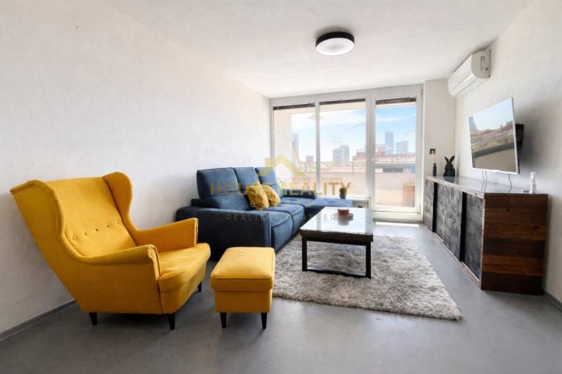 Nappali egy háromszobás lakásban sárga fotellel, kék kanapéval és falra szerelt TV-vel.