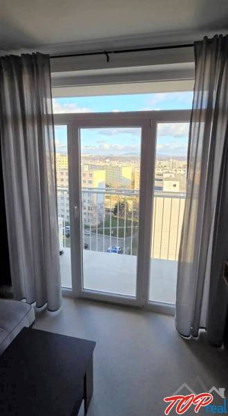 Garzonlakás kilátással az erkélyajtón keresztül, világos padló, függönyök.