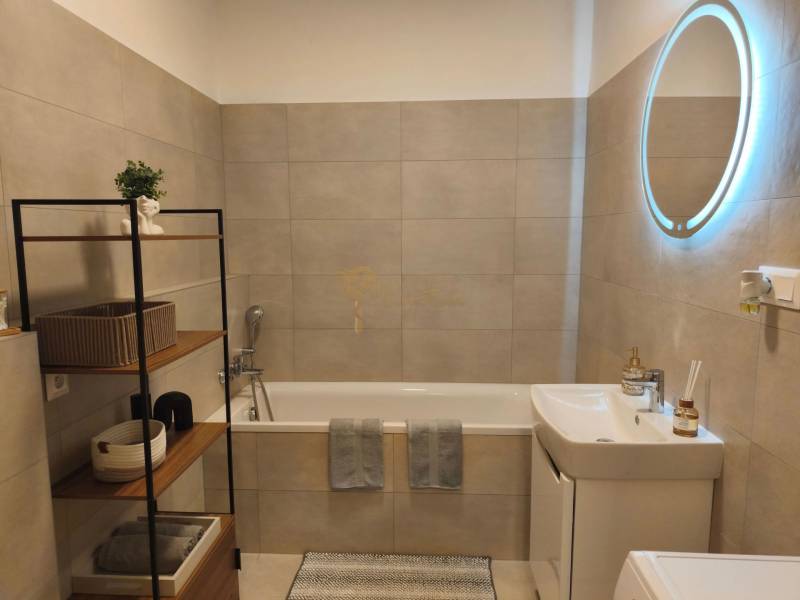 Kétszobás lakás fürdőszobája káddal, polcrendszerrel és világított tükörrel.