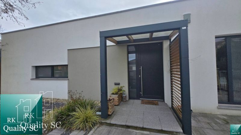 Családi ház Szencen minimalista homlokzattal, bejárati ajtóval és pergolával.