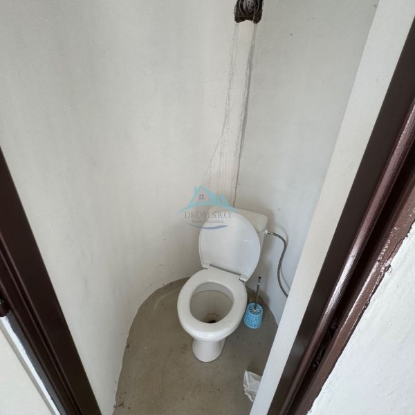 Egyszerű WC a házikóban, burkolat nélküli falakkal és műanyag WC-vel.