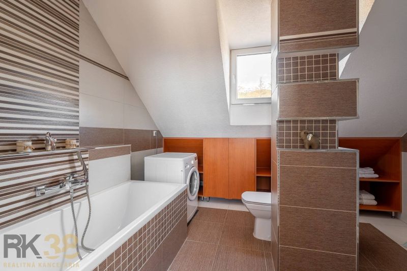 Tetőtéri fürdőszoba káddal, mosógéppel és barna csempével egy négy szobás lakásban.