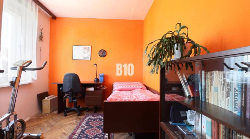 Szoba egy háromszobás lakásban berendezéssel: ágy, asztal, polc és szobabicikli.