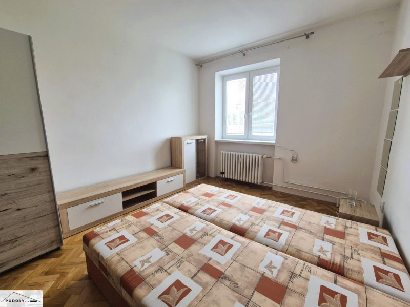 Szoba egy háromszobás lakásban, kétszemélyes ággyal és fa mintázatú padlóval.