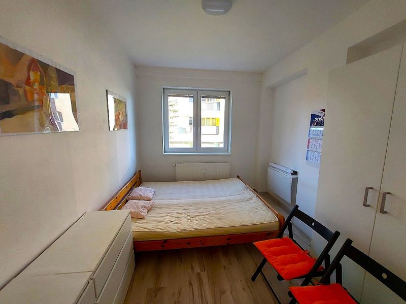 Garzonlakás ággyal, fehér szekrénnyel és fa mintázatú padlóval, két összecsukható székkel.