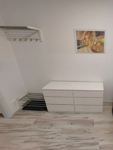 Garzonlakás fa hatású padlóval, fehér bútorokkal és színes festménnyel a falon.