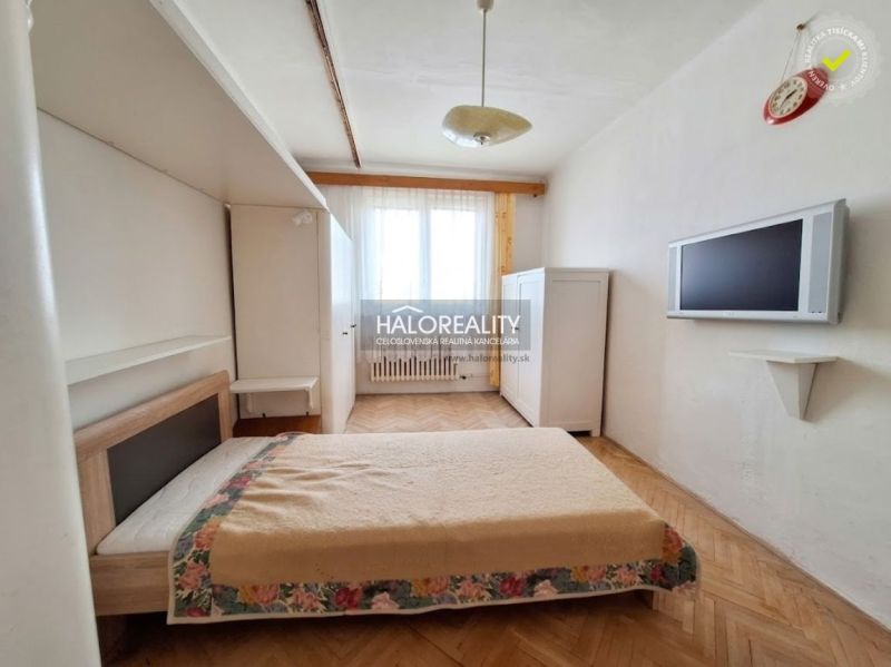 Hálószoba egy háromszobás lakásban ággyal, szekrénnyel, televízióval és fa mintázatú padlóval.