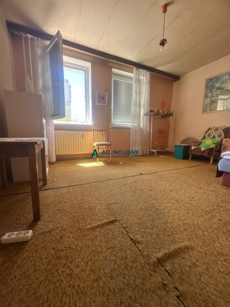 Szoba egy kétszobás lakásban szőnyeggel, nyitott ablakkal és egyszerű bútorokkal.