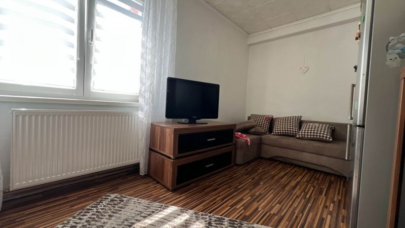 Nappali szoba fa hatású padlóval, kanapéval, televízióval és radiátorral egy háromszobás lakásban.