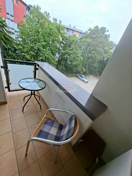 Garzonlakás erkélye kilátással a zöldre és parkolóra, szoba asztallal és székkel.