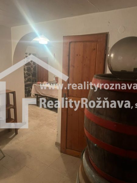 A kunyhó boltja egy fahordóval és kőfallal, a háttérben egy asztal látható.