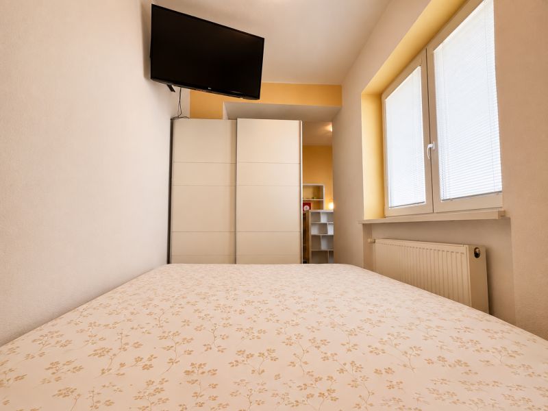 Hálószoba egy egyszobás lakásban, televízióval, szekrénnyel és redőnyös ablakkal.