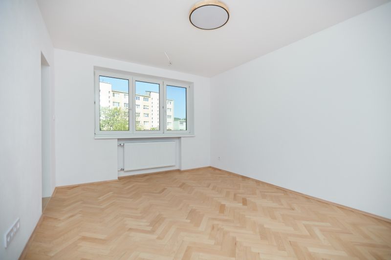 Szoba egy háromszobás lakásban fehér falakkal és fa mintázatú padlóval.