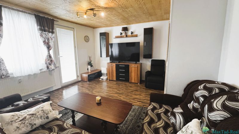 Nappali egy négy szobás lakásban, fa mintázatú padlóval és falra szerelt televízióval.