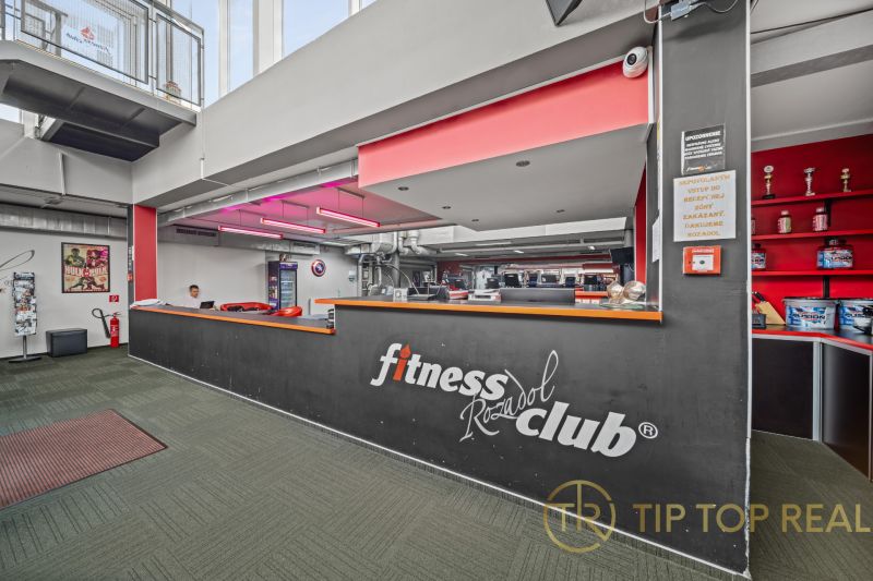 Egy fitnesz klub recepciója felirattal, piros világítással és dekoratív trófeákkal zöld árnyalatú padlón.