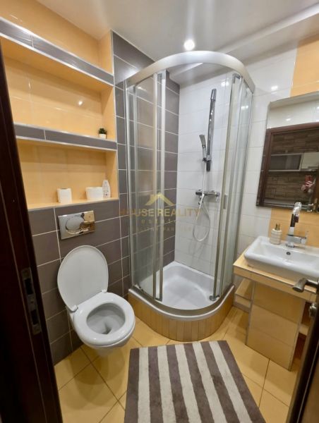 Fürdőszoba WC-vel, zuhanyzóval, mosdóval és polcokkal egy egyszobás lakásban.