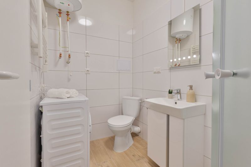 Fürdőszoba garzonlakásban mosógéppel, WC-vel, mosdóval és bojlerrel, fa mintázatú padlóval.