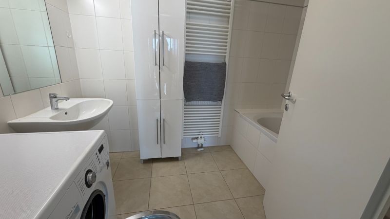 Fürdőszoba egy háromszobás lakásban mosógéppel, mosdóval és káddal világos csempéken.