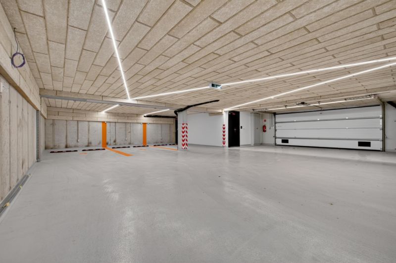 Beton mennyezetű és padlójú parkológarázs, lineáris lámpatestekkel megvilágítva.