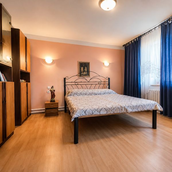 Hálószoba fém ággyal, fa mintázatú padlóval, bútorokkal és függönyökkel egy családi házban.