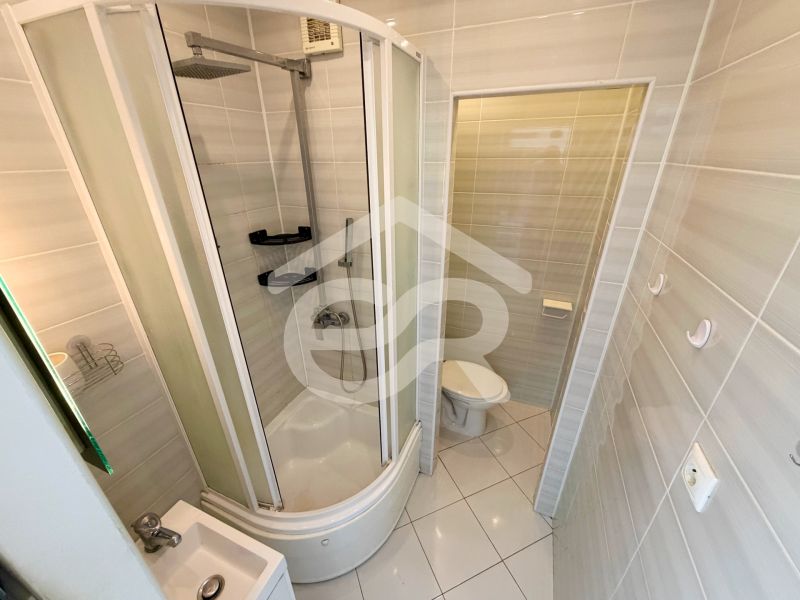 Fürdőszoba zuhanyzóval és WC-vel egy egyszobás lakásban, világos csempékkel.